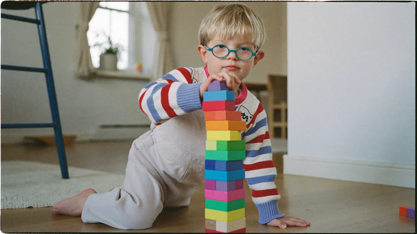 Ein kleiner Junge mit runder Brille baut auf dem Boden einen Turm aus bunten Bauklötzen.