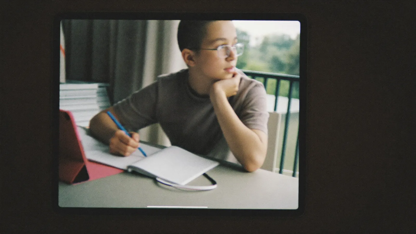 Ein Junge mit Brille sitzt nachdenklich an einem Tisch mit Heft und Tablet und schaut aus dem Fenster.