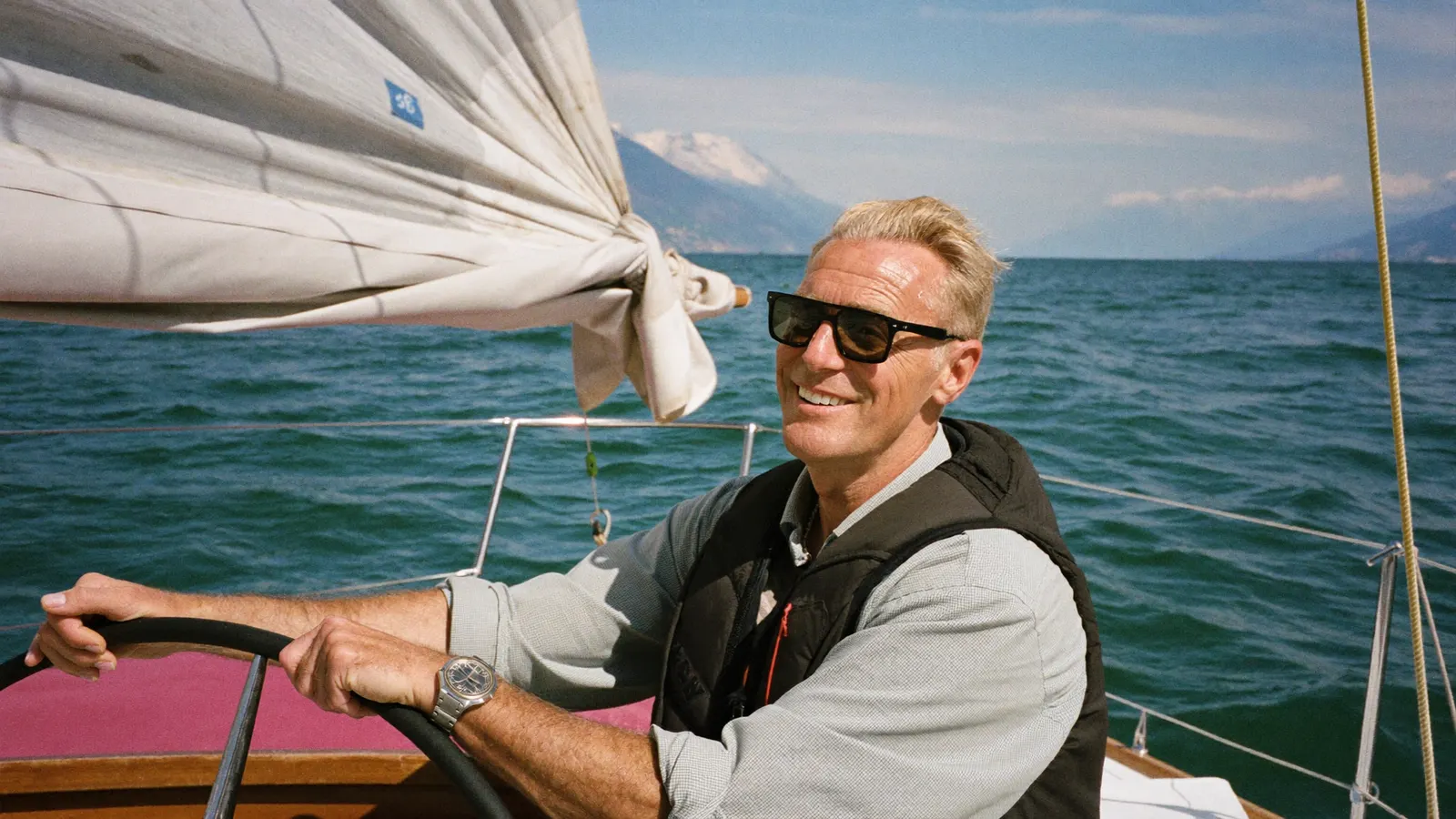 Ein Mann mit Sonnenbrille steuert lächelnd ein Segelboot auf einem See, im Hintergrund schneebedeckte Berge.