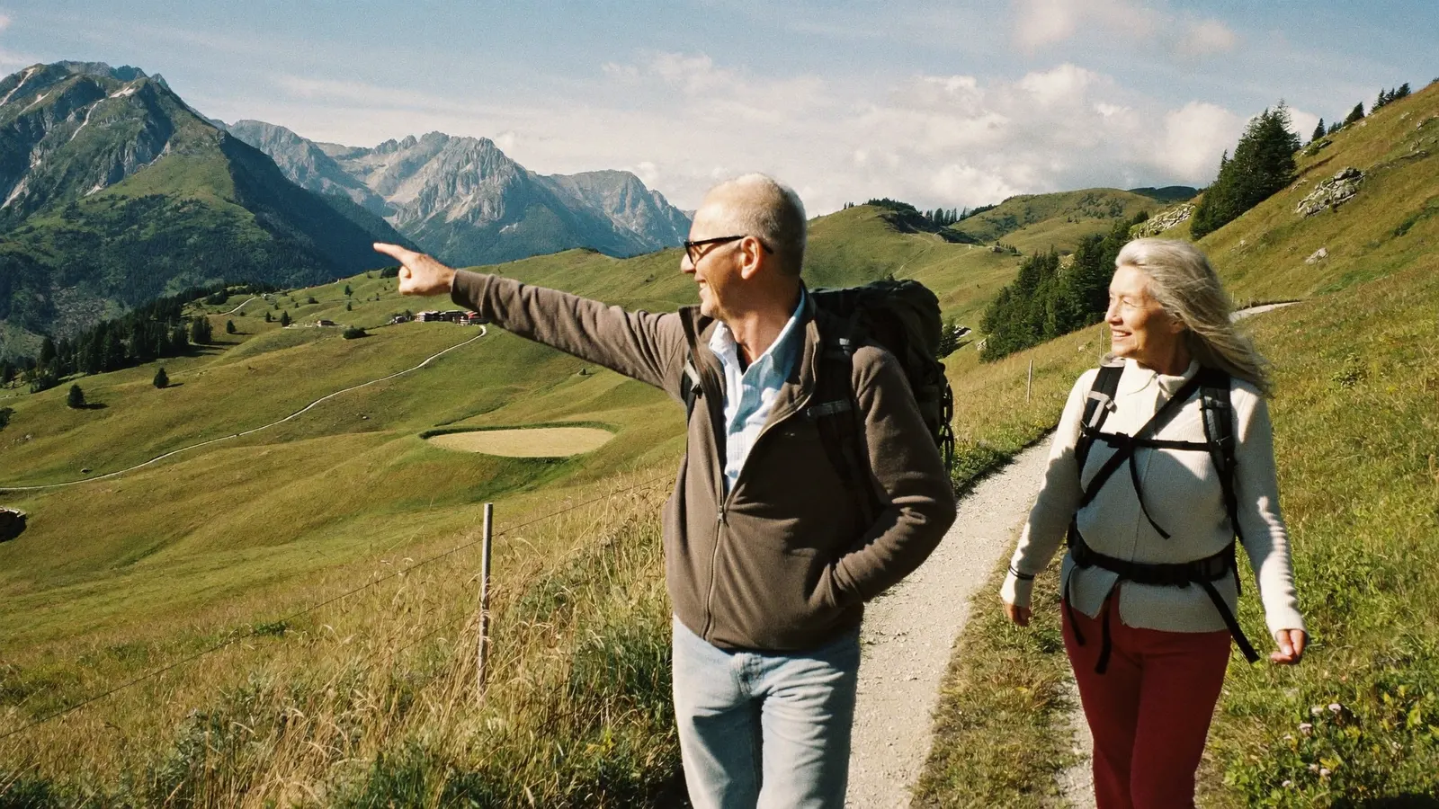 Ein älteres Paar wandert auf einem Bergweg über grüne Almwiesen, der Mann zeigt in die Ferne.